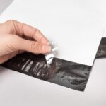 Kobieca dłoń odklejająca zabezpieczenie z paska kleju na białym foliopaku kurierskim