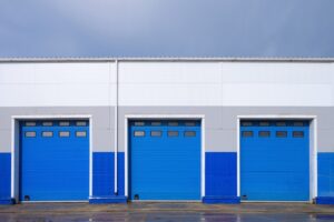 Trzy niebieskie bramy przemysłowe segmentowe z przeszkleniami w górnej części konstrukcji